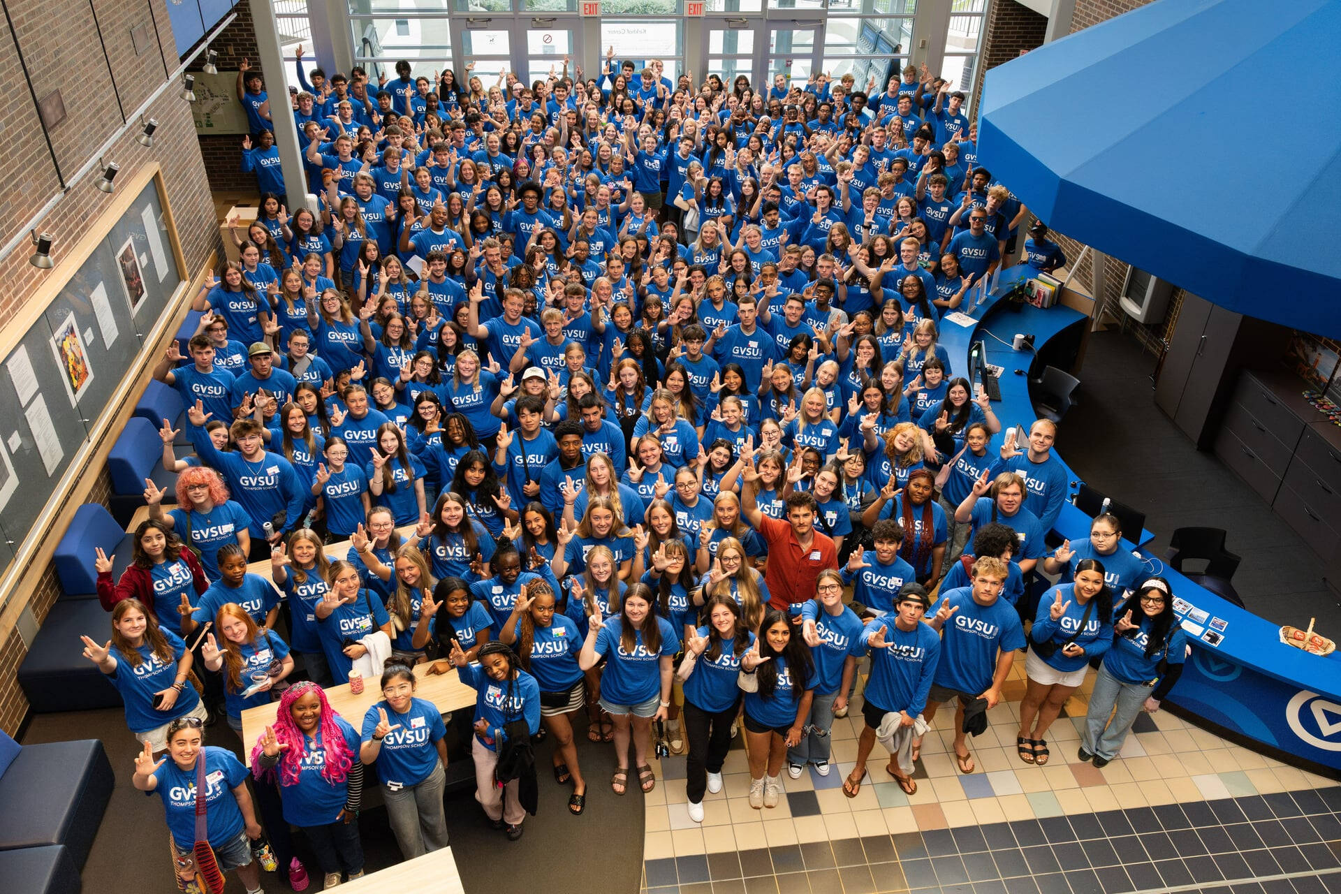 large group of Thompson Scholars in lobby of Kirkhof Center, many holding up hands in "L" sign, most in blue t-shirts